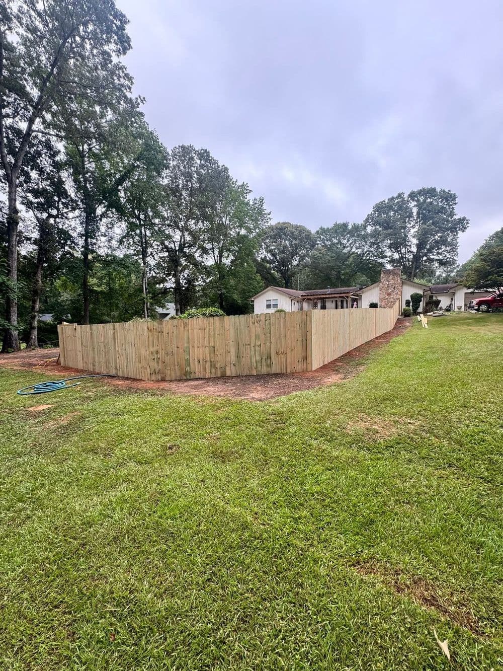 Wooden fence enclosing a yard with trees and a house in the background on a cloudy day.