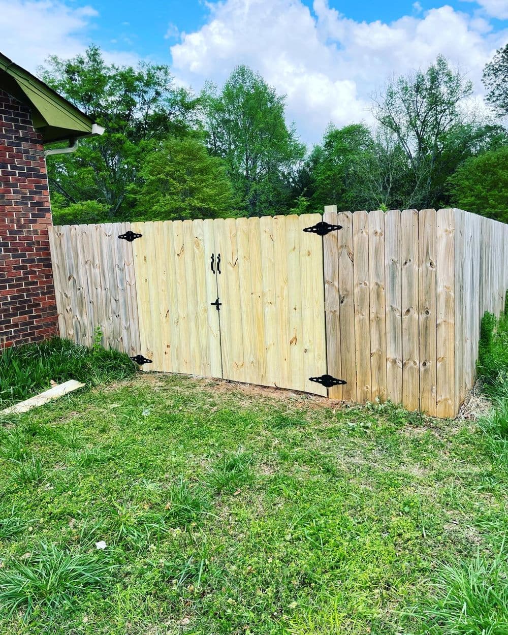 Wooden fence gate with black hardware in a grassy yard under a blue sky.