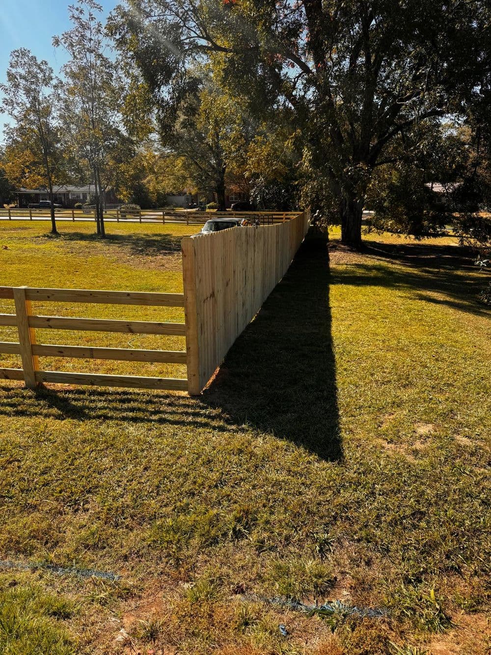 Wooden fence along a grassy yard, casting a shadow, surrounded by trees and open space.