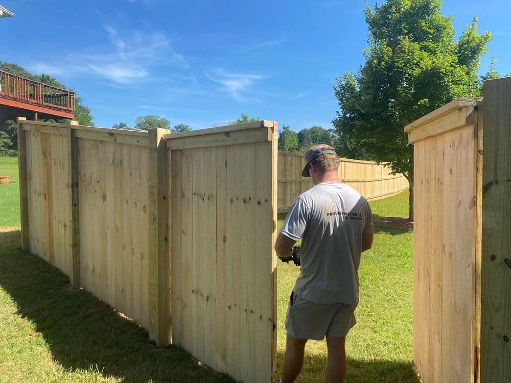 Man installing a wooden fence in a backyard under a clear blue sky.