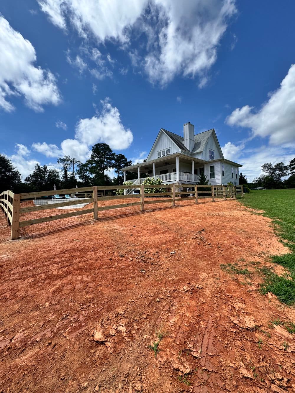 White farmhouse with a porch, fenced yard, and blue sky on a red dirt landscape.