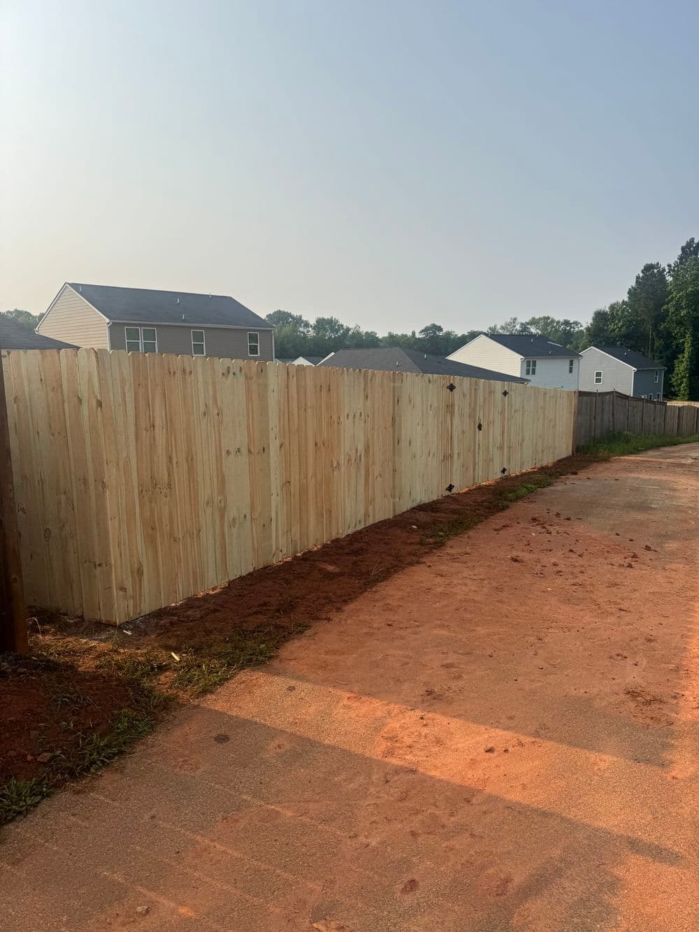 New wooden fence along dirt pathway with houses in the background on a sunny day.