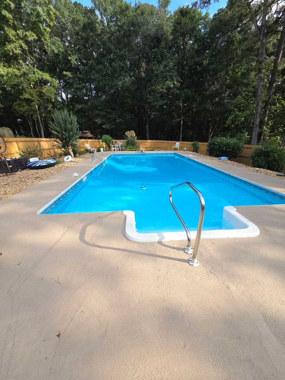 Backyard swimming pool surrounded by trees and landscaping, featuring a clear blue water.