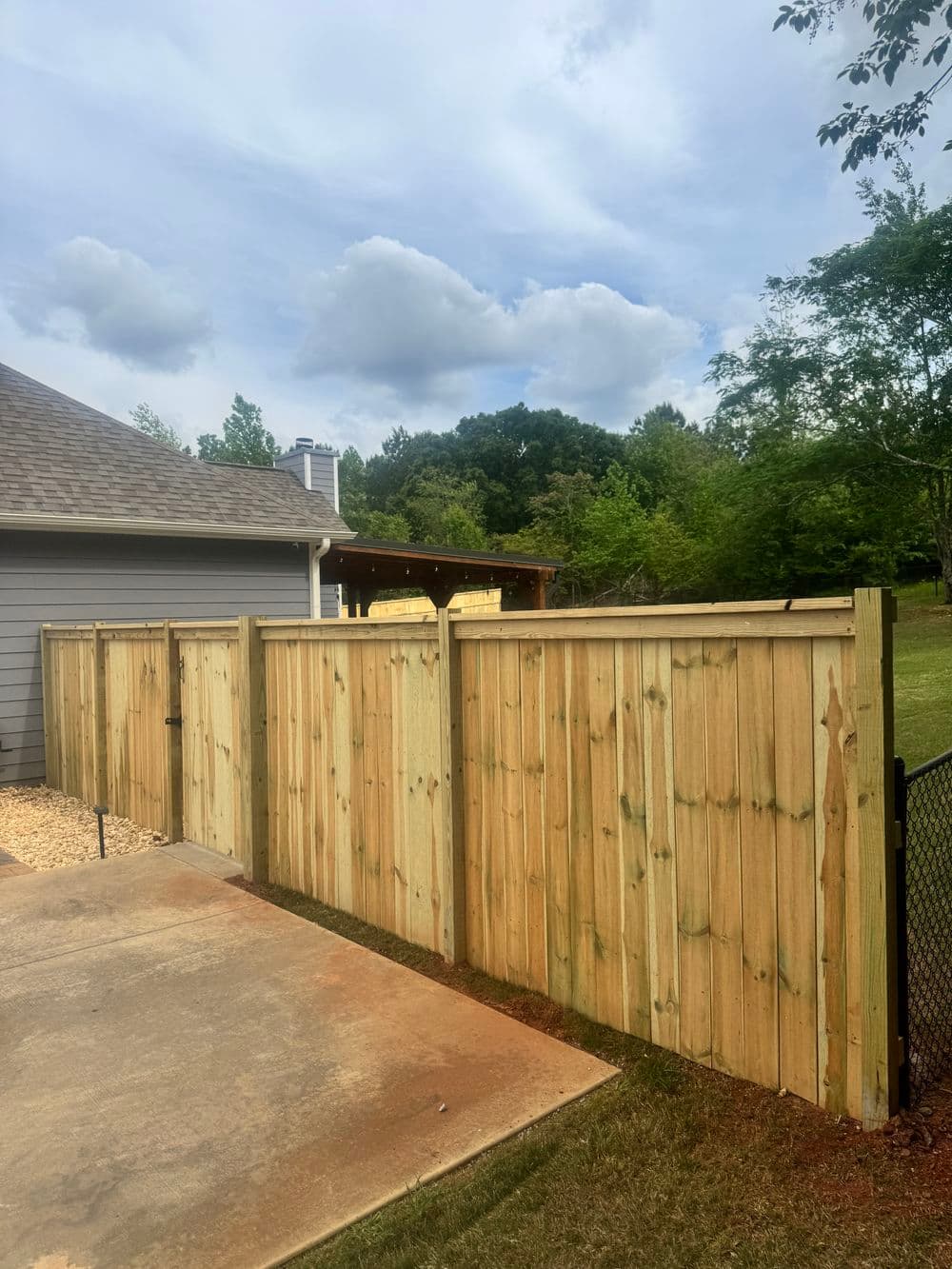 Wooden fence enclosing backyard with concrete pathway and lush greenery in the background.
