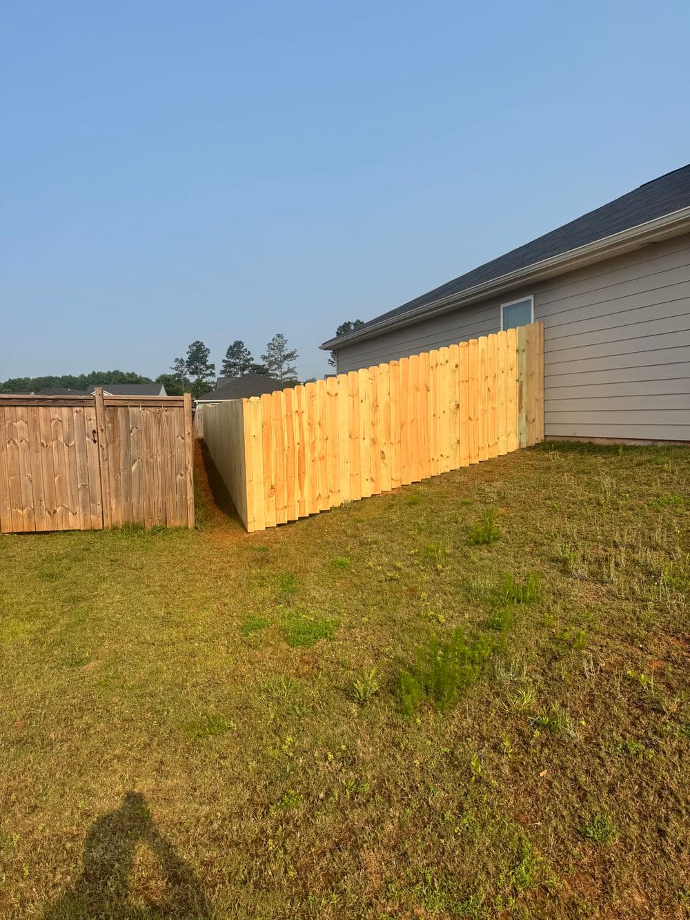 New wooden fence installed on a grassy slope beside a house under clear blue skies.