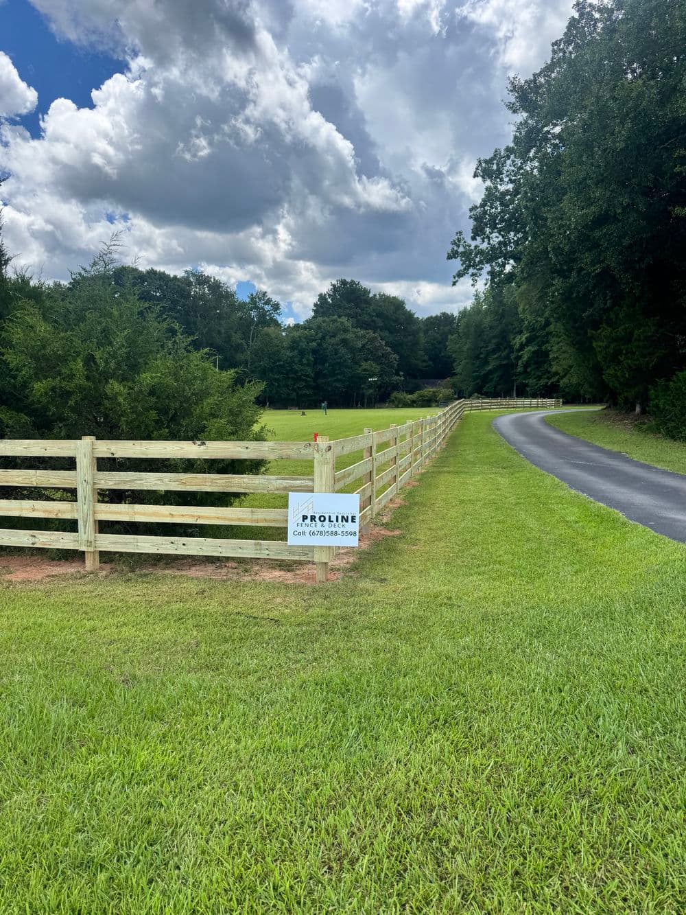 Wooden fence along a grassy path with a Proline sign and trees under a cloudy sky.