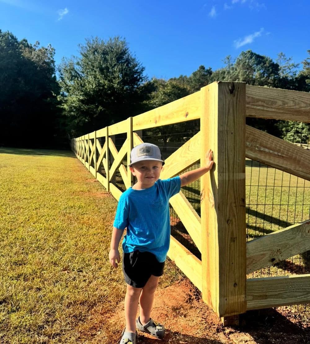 Young boy in a blue shirt standing by a wooden fence on a sunny day.