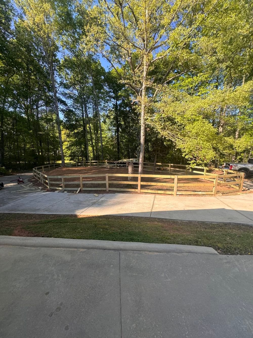 Wooden fenced area surrounded by trees, with a paved driveway in the foreground.