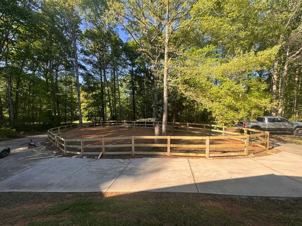 Circular fenced area with a central tree in a wooded landscape and nearby parked vehicles.