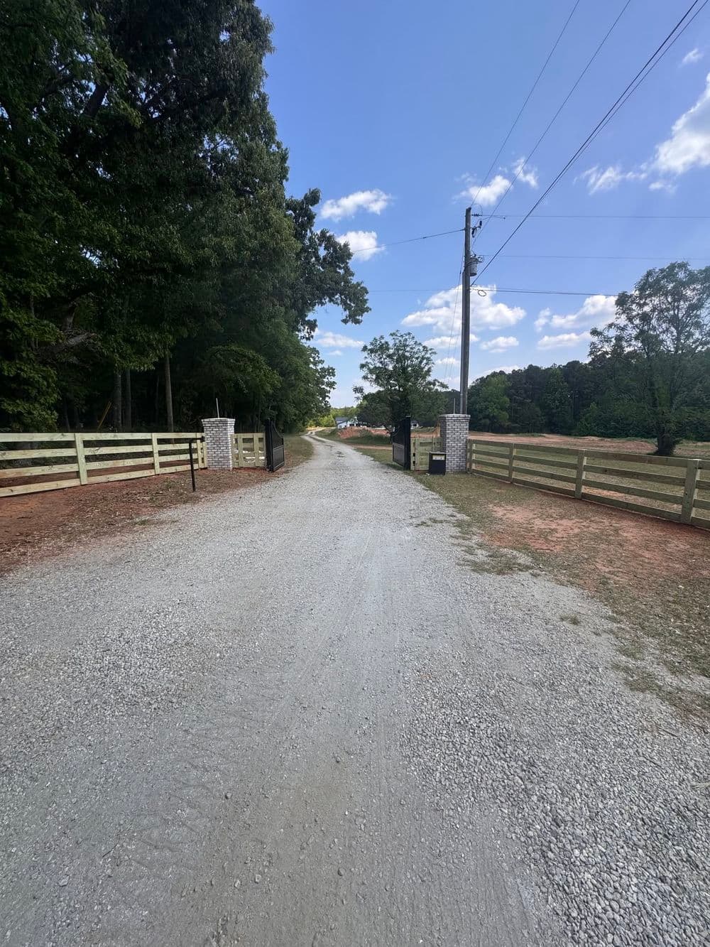 Gravel road leading to a gated property surrounded by trees and clear blue sky.